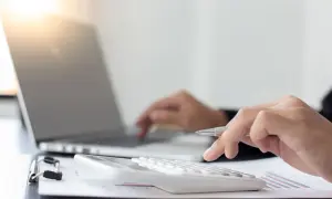 Dłoń z długopisem naciska klawisze kalkulatora, obok laptopa. Obliczenia podatku rolnego.