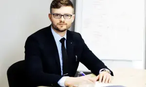 Mężczyzna w garniturze i okularach siedzi przy biurku, pisząc coś na kartce. Obok stoi filiżanka kawy.