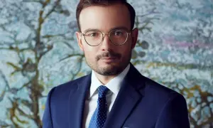 Mężczyzna w granatowym garniturze, białej koszuli i niebieskim krawacie w kropki, w okularach, na tle abstrakcyjnego obrazu.