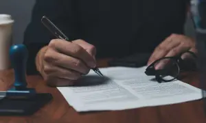 Dłoń podpisuje dokument piórem. Obok leżą okulary i pieczęć, gotowa do złożenia **żelazny** podpis.