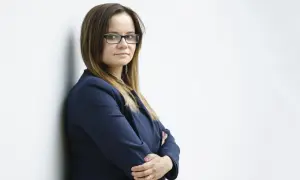 Kobieta w okularach i granatowej marynarce stoi oparta o ścianę, ze skrzyżowanymi rękami.
