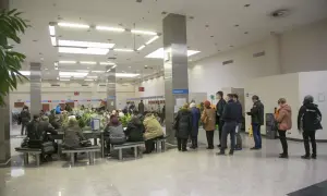 Ludzie czekają w kolejce do okienka, aby opłacić opłatę skarbową. W tle widać numery stanowisk i rośliny.