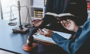 Dwie osoby w garniturach rozmawiają przy biurku z wagą sprawiedliwości, młotkiem i dokumentami.