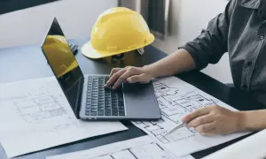 Inżynier analizuje plany budowlane, obliczając powierzchnię zabudowy na laptopie. Obok leży żółty kask.