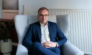 Mężczyzna w garniturze siedzi w fotelu, w tle kaloryfer i roślina.