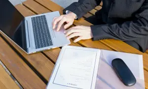 Biznesmen pracuje na laptopie, obok dokumenty i etui na okulary. Szykuje wpis do księgi wieczystej.