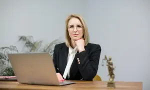 Kobieta w okularach i żakiecie siedzi przy biurku z laptopem i Statuą Temidy.