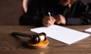 Młotek sędziowski na drewnianym stole, obok dokumentów. W tle osoba pisząca, być może oskarżyciel posiłkowy przygotowujący swoje pismo.