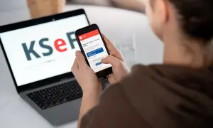 Osoba korzysta z telefonu z aplikacją KSeF, gdzie widnieje informacja o fakturze ustrukturyzowanej. Obok laptop z logo KSeF.