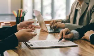 Dłonie biznesmenów przy biurku, jeden wskazuje na dokument, drugi pisze na laptopie.