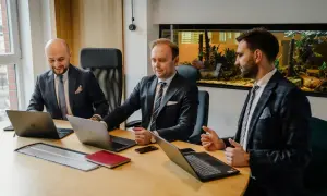 Trzech mężczyzn w garniturach przy drewnianym stole z laptopami i akwariem w tle.