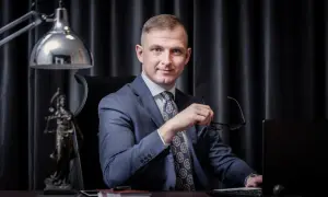 Mężczyzna w garniturze siedzi przy biurku, trzymając okulary. Obok statua Temidy.