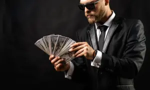 Elegancki mężczyzna w garniturze i okularach przeciwsłonecznych liczy banknoty. Wygląda jak windykator, który właśnie odzyskał dług.