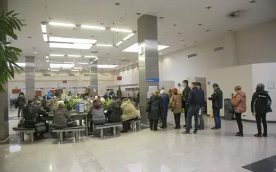 Ludzie czekają w kolejce do okienka, aby opłacić opłatę skarbową. W tle widać numery stanowisk i rośliny.