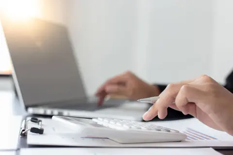 Dłoń z długopisem naciska klawisze kalkulatora, obok laptopa. Obliczenia podatku rolnego.