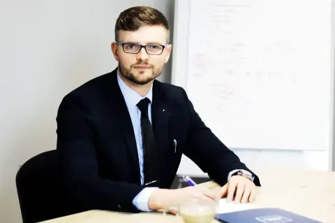 Mężczyzna w garniturze i okularach siedzi przy biurku, pisząc coś na kartce. Obok stoi filiżanka kawy.