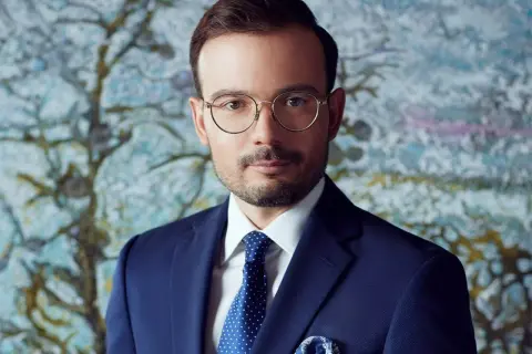 Mężczyzna w granatowym garniturze, białej koszuli i niebieskim krawacie w kropki, w okularach, na tle abstrakcyjnego obrazu.