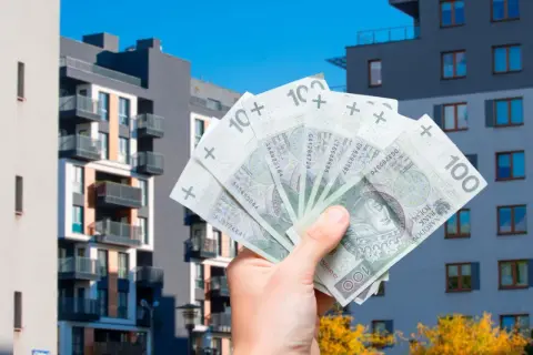 Dłoń trzyma banknoty 100 zł na tle nowoczesnych bloków mieszkalnych. To symbol ulgi mieszkaniowej i możliwości zakupu własnego M.