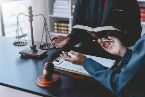 Dwie osoby w garniturach rozmawiają przy biurku z wagą sprawiedliwości, młotkiem i dokumentami.