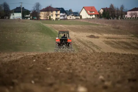 Czerwony traktor pracuje na polu, przygotowując ziemię do nowego sezonu. Odrolnienie działki trwa w najlepsze, a w tle widać domy.