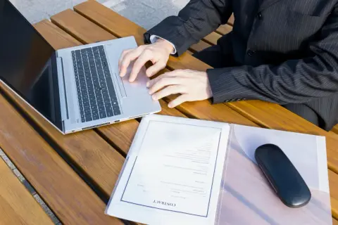 Biznesmen pracuje na laptopie, obok dokumenty i etui na okulary. Szykuje wpis do księgi wieczystej.