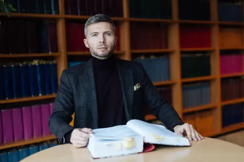 Mężczyzna w garniturze siedzi przy stole z otwartą księgą, otoczony regałami z książkami. Zastanawia się, ile bierze adwokat za reprezentowanie w sądzie.