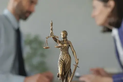 Figurka Temidy z wagą i mieczem na tle niewyraźnych postaci prawników.