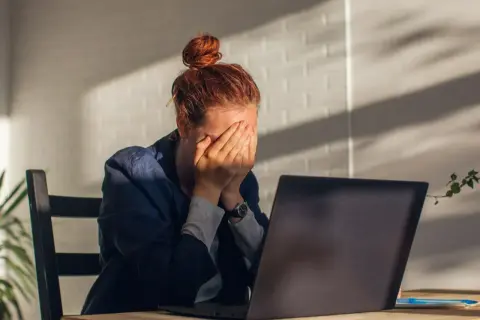 Kobieta z rudymi włosami, ukrywająca twarz w dłoniach, siedzi przy laptopie. Widać oznaki wypalenia zawodowego, być może potrzebne jest L4.