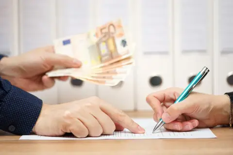 Dwie osoby przy biurku. Jedna podaje plik banknotów 50 euro, druga podpisuje dokument. To może być moment, kiedy należy się odprawa.