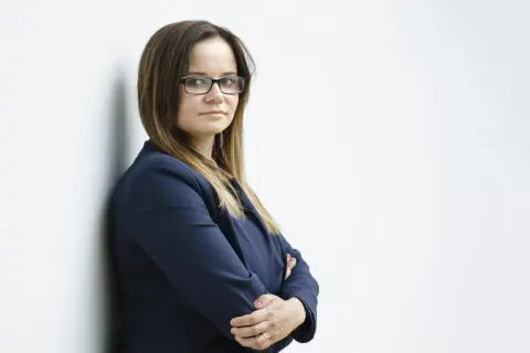 Kobieta w okularach i granatowej marynarce, oparta o białą ścianę.