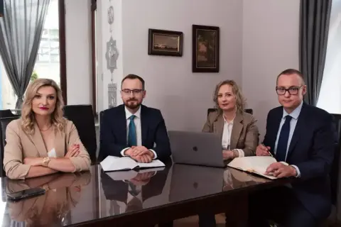 Cztery osoby w strojach biznesowych siedzą przy stole w sali konferencyjnej.