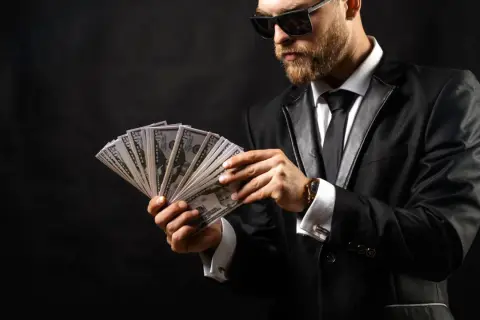 Elegancki mężczyzna w garniturze i okularach przeciwsłonecznych liczy banknoty. Wygląda jak windykator, który właśnie odzyskał dług.