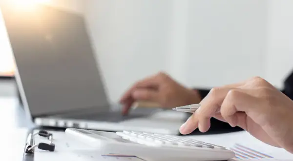 Dłoń z długopisem naciska klawisze kalkulatora, obok laptopa. Obliczenia podatku rolnego.