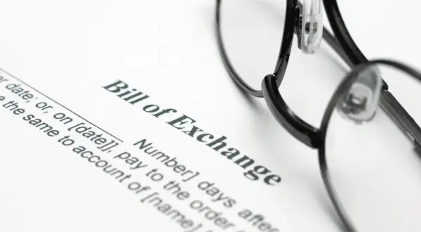 Okulary leżą na dokumencie "Bill of Exchange", czyli wekslu. Widoczne są fragmenty tekstu dotyczące daty i płatności.