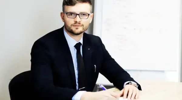 Mężczyzna w garniturze i okularach siedzi przy biurku, pisząc coś na kartce. Obok stoi filiżanka kawy.