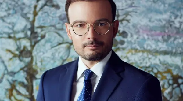 Mężczyzna w granatowym garniturze, białej koszuli i niebieskim krawacie w kropki, w okularach, na tle abstrakcyjnego obrazu.