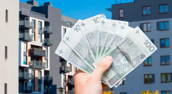 Dłoń trzyma banknoty 100 zł na tle nowoczesnych bloków mieszkalnych. To symbol ulgi mieszkaniowej i możliwości zakupu własnego M.