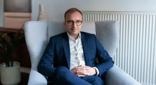 Mężczyzna w garniturze siedzi w fotelu, w tle kaloryfer i roślina.