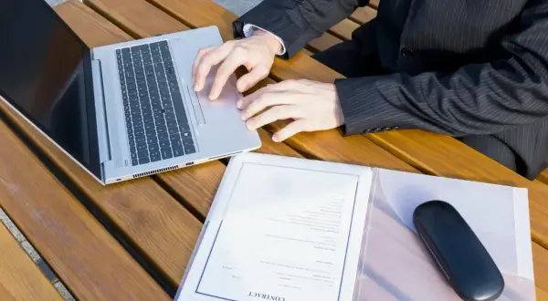 Biznesmen pracuje na laptopie, obok dokumenty i etui na okulary. Szykuje wpis do księgi wieczystej.