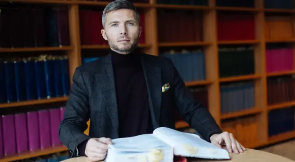 Mężczyzna w garniturze siedzi przy stole z otwartą księgą, otoczony regałami z książkami. Zastanawia się, ile bierze adwokat za reprezentowanie w sądzie.