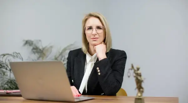 Kobieta w okularach i żakiecie siedzi przy biurku z laptopem i Statuą Temidy.