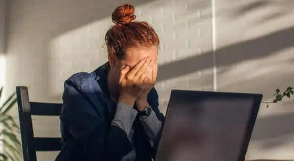Kobieta z rudymi włosami, ukrywająca twarz w dłoniach, siedzi przy laptopie. Widać oznaki wypalenia zawodowego, być może potrzebne jest L4.