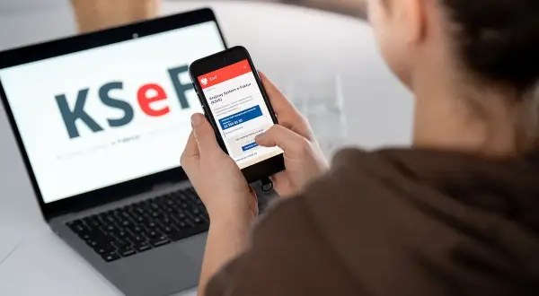Osoba korzysta z telefonu z aplikacją KSeF, gdzie widnieje informacja o fakturze ustrukturyzowanej. Obok laptop z logo KSeF.