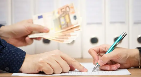Dwie osoby przy biurku. Jedna podaje plik banknotów 50 euro, druga podpisuje dokument. To może być moment, kiedy należy się odprawa.