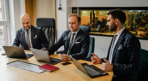 Trzech mężczyzn w garniturach przy drewnianym stole z laptopami i akwariem w tle.