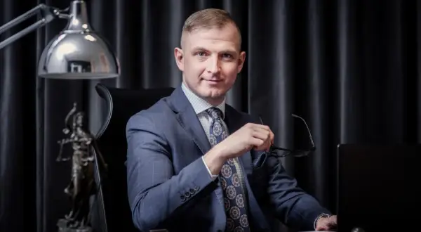 Mężczyzna w garniturze siedzi przy biurku, trzymając okulary. Obok statua Temidy.