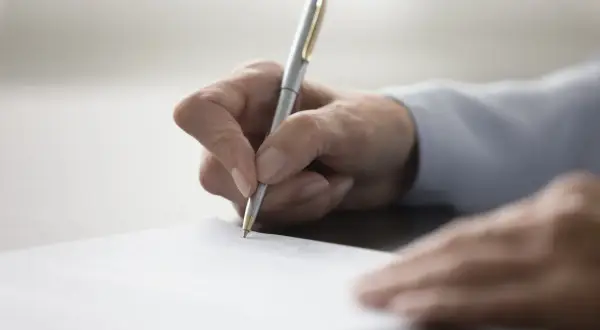 Dłoń pisze długopisem na papierze, przygotowując dokument. Dowiedz się, jak napisać testament.