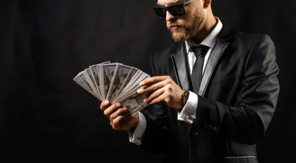 Elegancki mężczyzna w garniturze i okularach przeciwsłonecznych liczy banknoty. Wygląda jak windykator, który właśnie odzyskał dług.
