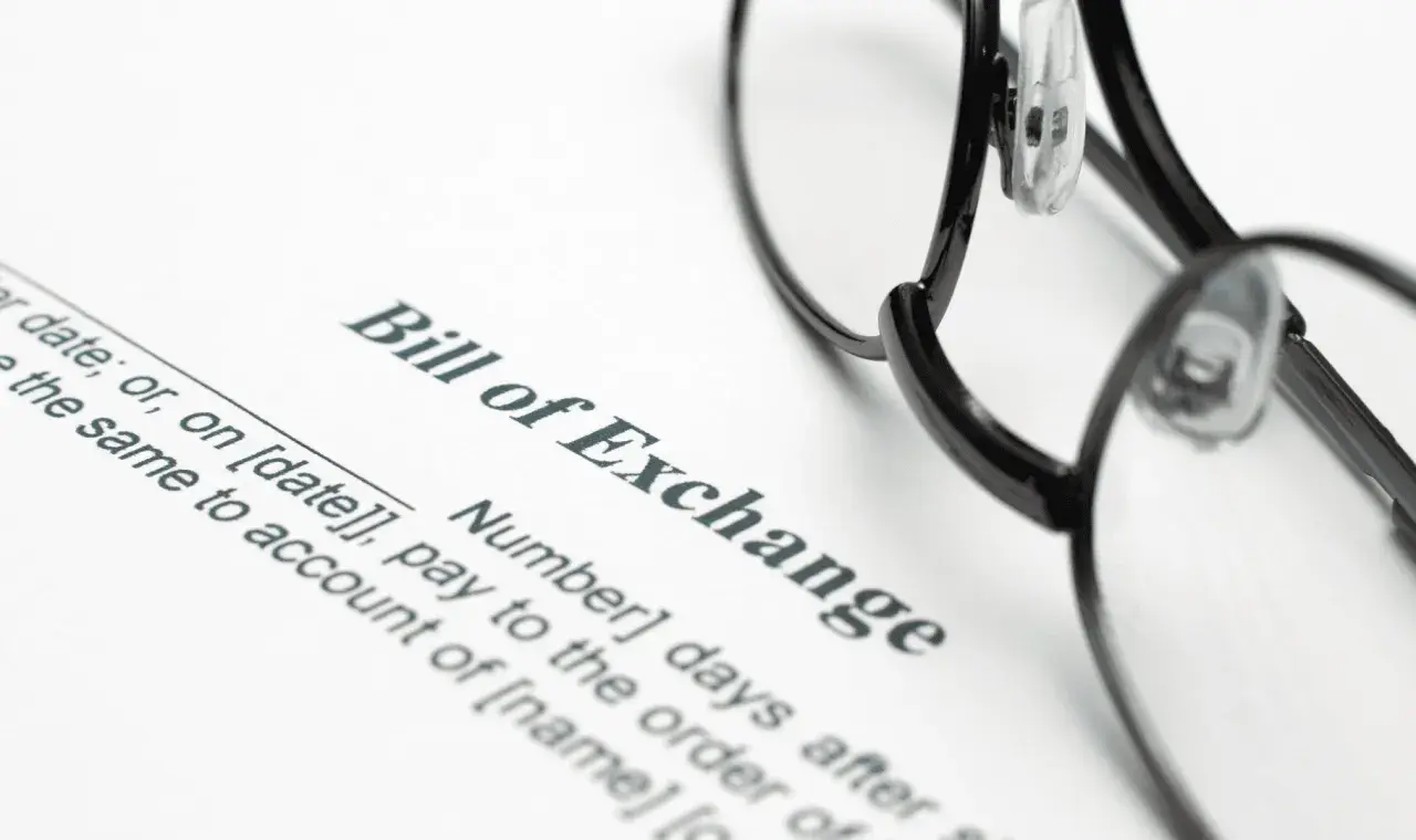 Okulary leżą na dokumencie "Bill of Exchange", czyli wekslu. Widoczne są fragmenty tekstu dotyczące daty i płatności.