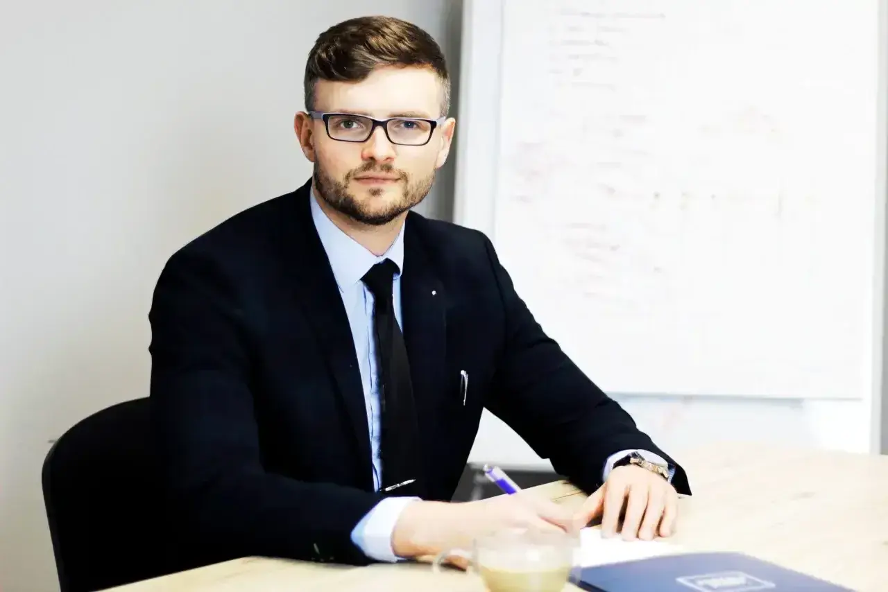 Mężczyzna w garniturze i okularach siedzi przy biurku, pisząc coś na kartce. Obok stoi filiżanka kawy.